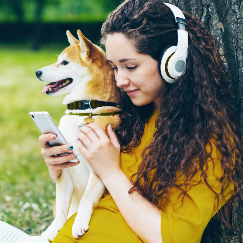 Frau mit Kopfhörern sitzt neben ihrem Hund auf einer Wiese und schaut auf ihr Smartphone